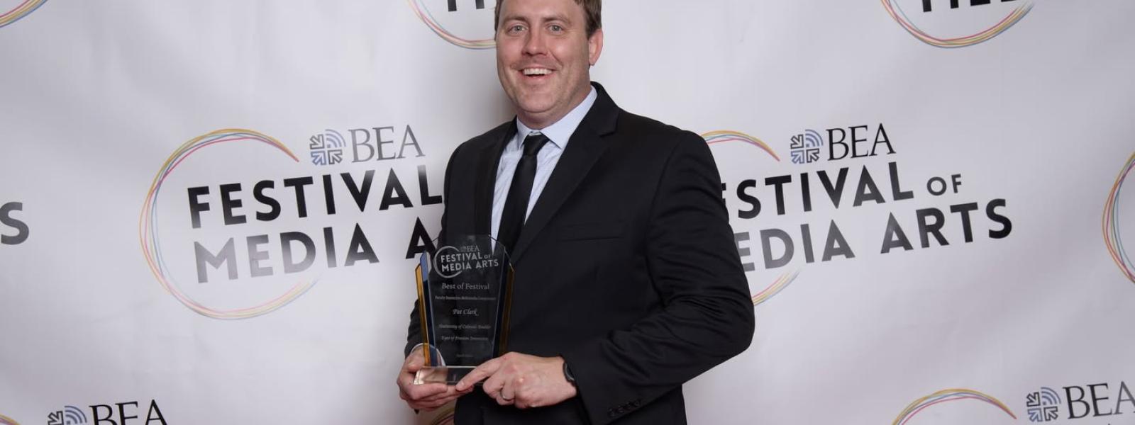 Pat Clark holds an award in his hand and smiles for a photo in front of a white backdrop with "Festival of Media Arts" written in black text repeated across the backdrop