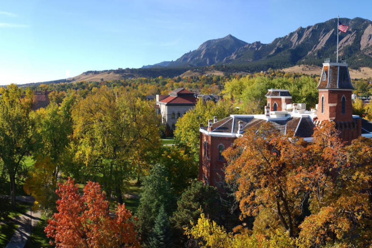 A panoramic image of the CU Boulder campus, with Old Main building in the foreground surrounded by lots of foliage, and the Flatiron mountains and a blue sky in the background.